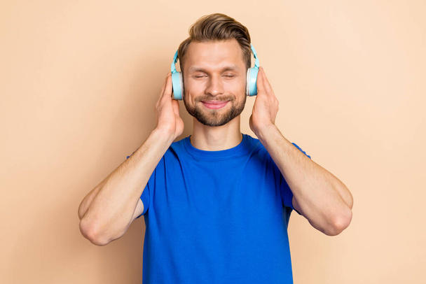 Photo of young guy good mood dreamy hands touch earphones sound isolated over beige color background. - Foto, immagini