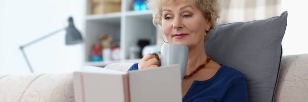 Ritratto di vecchia signora che legge un libro e beve una tazza di caffè comodamente sul divano di casa. Attraente donna anziana rilassante nel fine settimana. Concetto di tempo di ricambio - Foto, immagini
