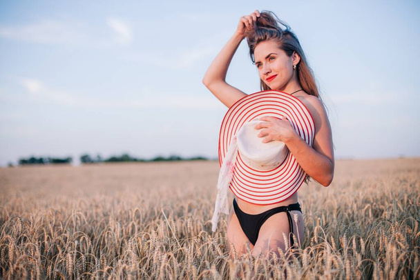 A young, slender, in black panties in a field of wheat covers her chest with a straw hat. High quality photo - Foto, imagen