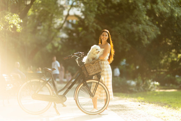 Pretty young woman with white bichon frise dog in the basket of electric bike - Foto, imagen