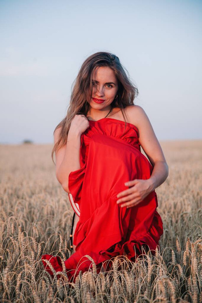 A young, slender girl with loose hair in a field of wheat covers her naked body with a red dress. High quality photo - Fotoğraf, Görsel