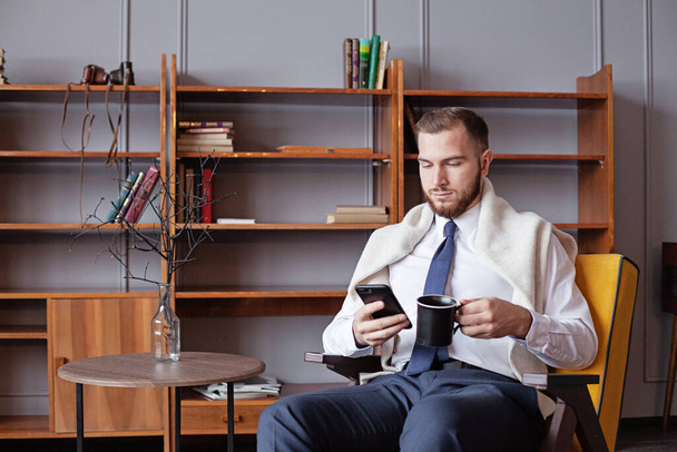 Retrato de un joven hombre de negocios usando un teléfono inteligente y sosteniendo una taza de café - Foto, imagen