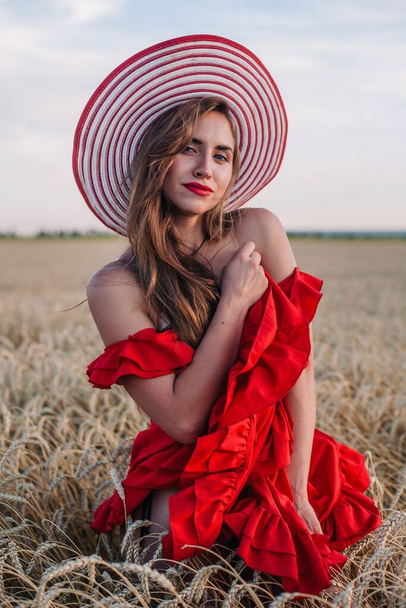 A young, slender, sexy girl in a bright red dress and hat stands in a field of wheat at sunset. High quality photo - Zdjęcie, obraz