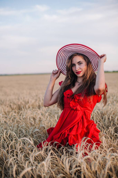 A young, slender, sexy girl in a bright red dress and hat stands in a field of wheat at sunset. High quality photo - Foto, Imagem