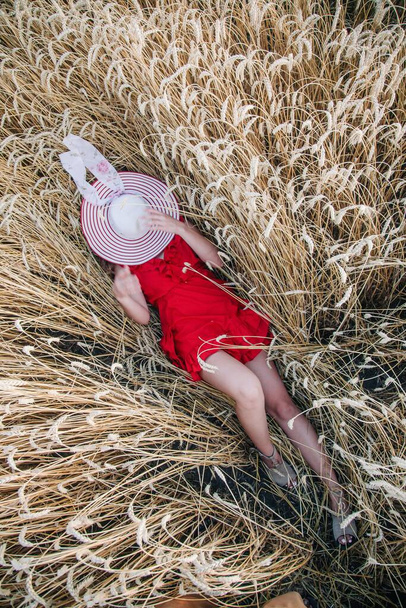 Uma menina jovem, esbelta e sexy em um vestido vermelho brilhante e chapéu está em um campo de trigo. Foto de alta qualidade - Foto, Imagem