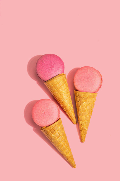 Ice cream waffle cones with macarons on pink background. Sweet dessert, summer concept. Trendy hard light, dark shadow, top view - 写真・画像