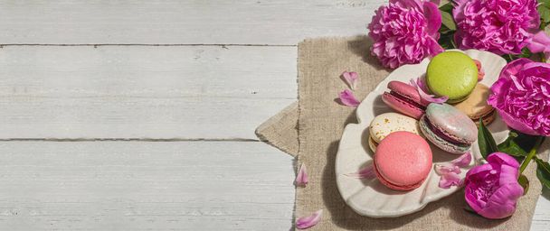 Macarons avec un bouquet de pivoines sur fond de bois blanc. Dessert doux, couleurs colorées et pastel, concept d'été. Romantique style rustique, carte festive, lumière dure, ombre sombre, format bannière - Photo, image