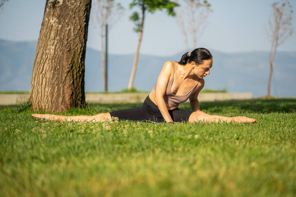 Una donna in reggiseno sportivo blu e nero breve seduta su tappetino yoga facendo la cavitazione con la mano insieme al petto sul pavimento in erba e giardino sfondo sfocato. - Foto, immagini
