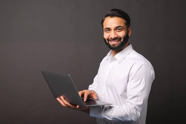 Indian male entrepreneur carrying laptop isolated on black, bearded businessman using laptop, typing, receiving emails, using new computer app, looking at the camera and smiles - Valokuva, kuva