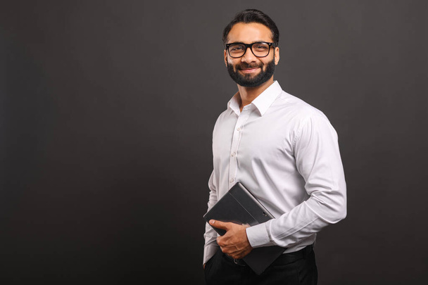 Indian male entrepreneur in formal white shirt and glasses carrying laptop isolated on black, bearded businessman using laptop, using new computer app, looking at the camera and smiles - Фото, зображення