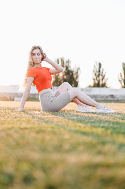 Frau in rotem Sporttop sitzt auf grünem Gras im Sonnenstrahl und schaut weg. Mädchen setzt sich nach dem Training zur Ruhe - Foto, Bild