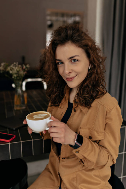 Stylish european woman with curls wearing brown shirt holding cup with coffee and smiling at camera. Indoor photo of lovely girl enjoying breakfast in cafe.  - 写真・画像
