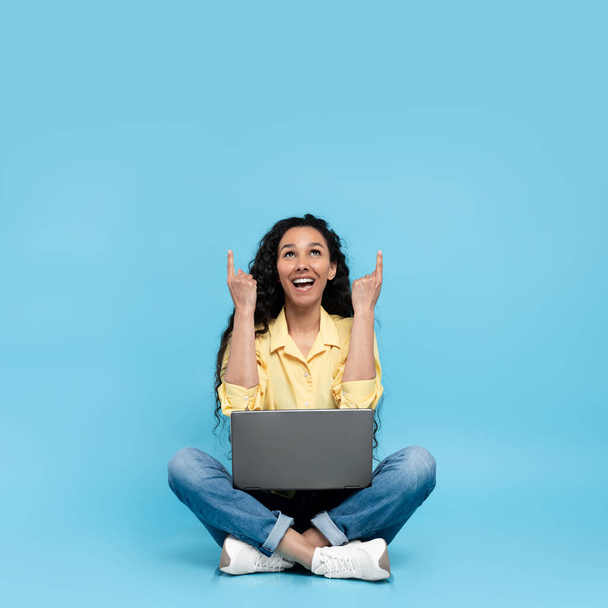 幸せな若い女性がオンラインで作業し、クロスレッグに座ってラップトップを使用し、青いスタジオの背景にある空のスペースを指しています。楽しいです千年の女性とともにPC提供場所のための広告 - 写真・画像