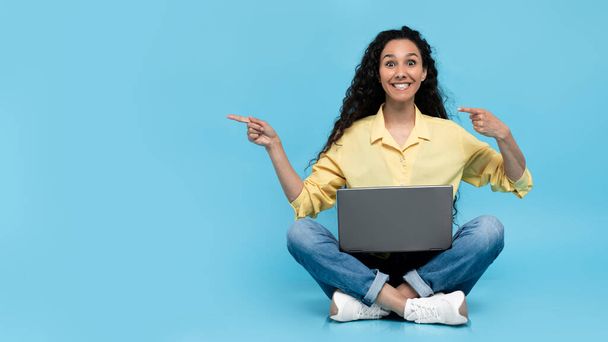 Portré vidám Y-generációs hölgy alkalmi viselet segítségével laptop számítógép az online munka, az oktatás vagy a kommunikáció, rámutatva az üres tér kék stúdió háttér, banner design - Fotó, kép