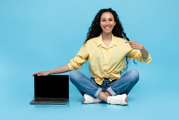 Full length of cheery young woman pointing at laptop computer with empty screen, προσφέροντας mockup για την ιστοσελίδα σε μπλε φόντο. Millennial θηλυκό επιδεικνύοντας σύγχρονο υπολογιστή, χώρος για διαφήμιση - Φωτογραφία, εικόνα