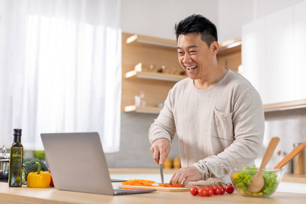 Wesoły mężczyzna w średnim wieku gotowanie obiad w domu, oglądanie receptury wideo w Internecie i cięcia warzyw, czytanie bloga żywności, za pomocą laptopa, wnętrze kuchni, przestrzeń do kopiowania. Gotowanie, kulinarne - Zdjęcie, obraz