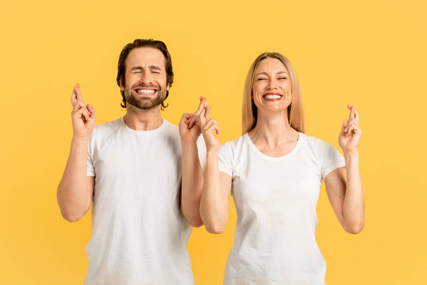 Cheerful millennial caucasian couple in white t-shirts with closed eyes, crossed fingers, make wish, dreaming isolated on yellow background. Hope, human emotions, ad and offer and facial expression - Photo, Image