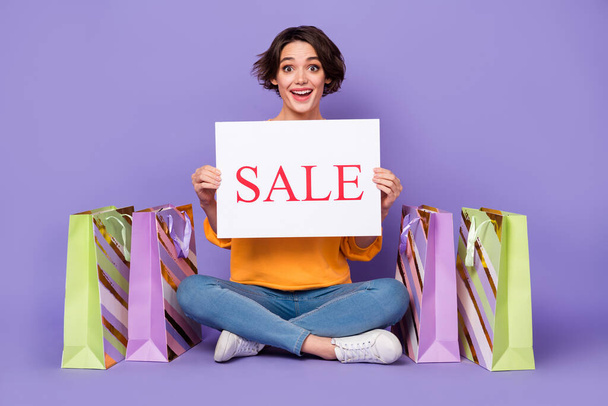 Portrait of attractive amazed cheerful girl holding sale board sitting lotus pose isolated over violet lilac color background. - Valokuva, kuva