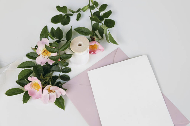 Summer wedding stationery mock-up scene with silk ribbon. Closeup of blank greeting card, invitation, pink envelope and blooming dog rose flowers isolated on white table background. Top view. - 写真・画像