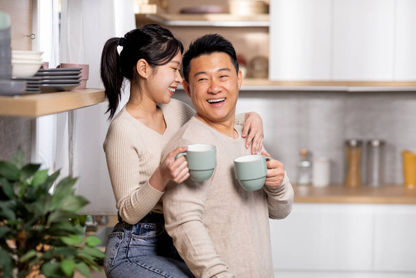 Portret wesołych koreańskich małżonków pijących herbatę w kuchni, ładna azjatycka kobieta siedząca na stole, przytulająca swojego śmiejącego się męża, ciesząca się razem czasem, widok z boku, przestrzeń na ksero. Rodzina, związki - Zdjęcie, obraz