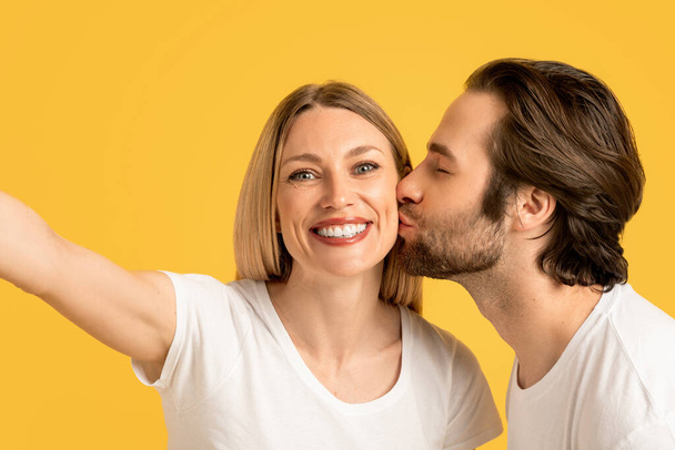 Happy millennial caucasian husband kissing wife on cheek in white t-shirt, lady make selfie, isolated on yellow background, close up. Photo for blog and social networks. Love, romance, relationship - Photo, Image