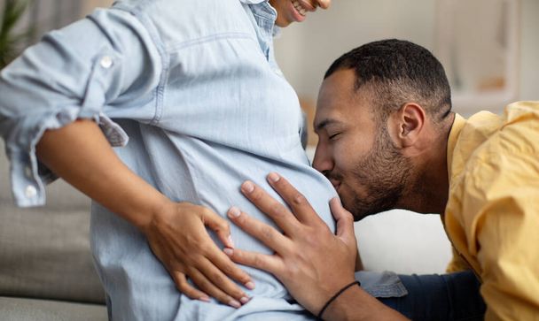 Terhesség és szülés. Vágott lövés az afro-amerikai férj csók feleségek hasa ül kanapén otthon. Családi várandós baba, szerelem és boldogság. Fogalom - Fotó, kép