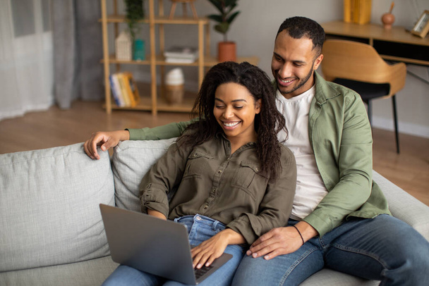 Happy African American Couple Using Laptop Computer Watching Movie Online And Websurfing Relaxing Together Sitting On Sofa At Home On Weekend. Technology And Gadgets Concept - Photo, image