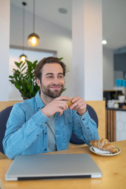 Time for yourself. Happy young bearded man holding cup sitting at table with laptop in cafe resting smiling to side - Foto, Bild
