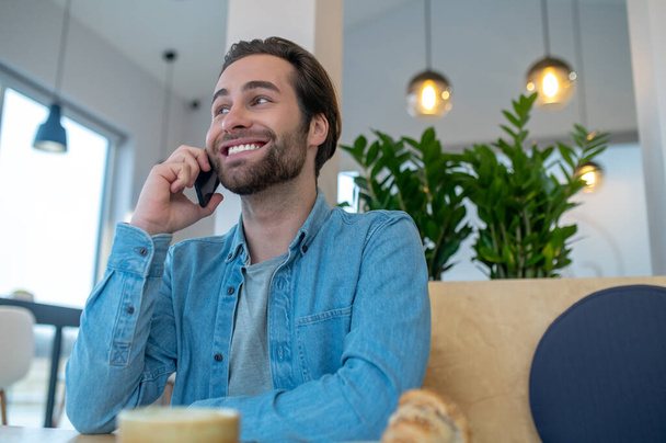 Phone call. Smiling young bearded man looking away talking on smartphone sitting indoors during day - Foto, Imagen