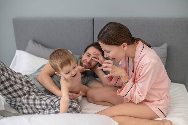 Playful mood. Emotional young woman in pajamas sitting playfully stretching hands to joyful child near dad reclining on bed in bedroom - Photo, Image