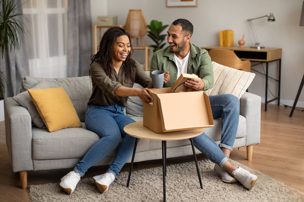 Радостный афро-американский муж и жена распаковывают картонную коробку, получая посылку, сидящую на диване в помещении. Пара празднуя успех онлайн покупки и доставка на дому - Фото, изображение