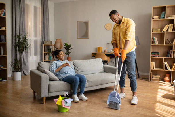Εγκυμοσύνη Αφρικανός Αμερικανός σύζυγος καθαρίζει το σπίτι ενώ η σύζυγος ξεκουράζεται και χρησιμοποιεί το κινητό τηλέφωνο κάθεται στον καναπέ στο σπίτι. Οικογενειακός τρόπος ζωής και οικιακή εργασία έννοια αναψυχής - Φωτογραφία, εικόνα