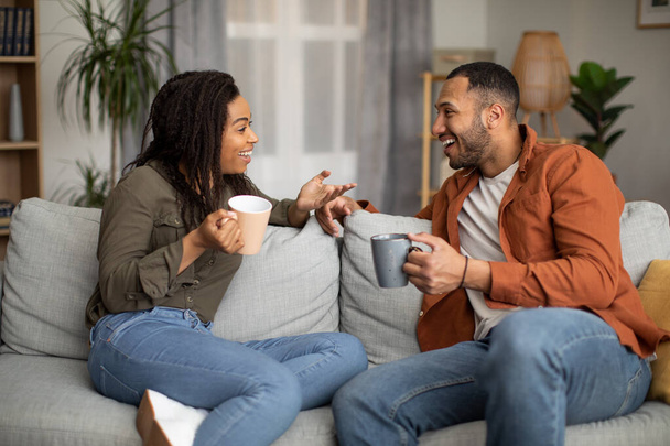 Iloiset mustat puolisot juovat kahvia ja puhuvat sohvalla kotona. Poikaystävä ja tyttöystävä Nauti keskustelusta Flirttailu Viikonloppu yhdessä. Suhde ja viestintä - Valokuva, kuva
