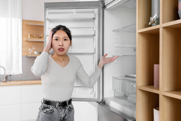 Megdöbbent fiatal koreai hölgy alkalmi áll nyitott üres hűtőszekrény és megérinti a fejét, az élelmiszer-hiány, a vásárlás szükségessége, szenved a pénzügyi másnaposság, míg világjárvány, másolás helyet - Fotó, kép