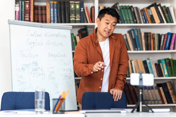 Asyalı erkek hoca, modern sınıftaki beyaz tahtanın yanında duran akıllı telefondan video görüşmesi yapıyor. İnternet Eğitimi ve Eğitim Günlüğü Kavramı - Fotoğraf, Görsel