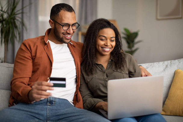 Happy African American para Zakupy online za pośrednictwem laptopa i karty kredytowej siedzi na kanapie w domu. Małżonkowie płacący za korzystanie z usługi bankowości internetowej na komputerze. Koncepcja handlu elektronicznego - Zdjęcie, obraz