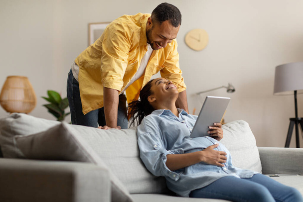 Cheerful Black Couple Using Digital Tablet Browsing Internet Sitting On Sofa At Home. Spouses Expecting Baby And Browsing Internet Via Computer On Weekend. Pregnancy Lifestyle, Technology - Φωτογραφία, εικόνα