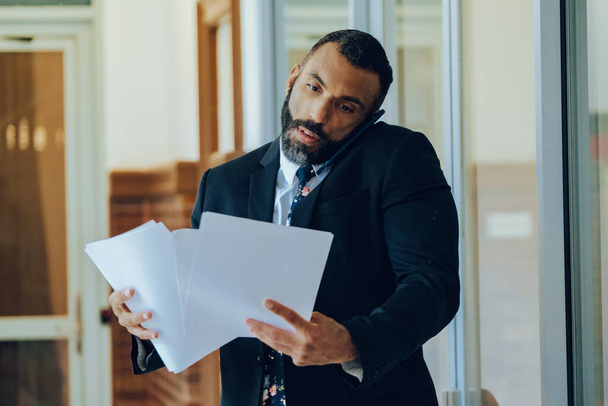 όμορφος Αφροαμερικάνος επιχειρηματίας με κοστούμι και γραβάτα μιλώντας σε smartphone και κρατώντας έγγραφα - Φωτογραφία, εικόνα