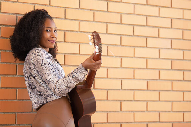 Smiling african american woman standing playing guitar looking at camera leaning against a brick wall. - Foto, imagen