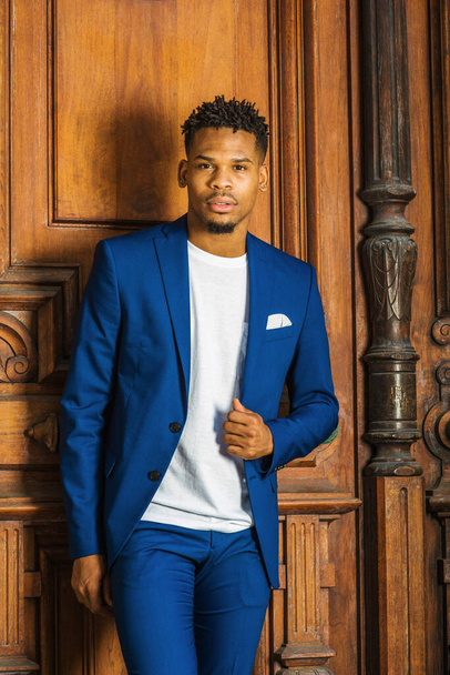 African American Businessman working in New York. Wearing blue suit, unbuttoned, white T shirt, black college student with little goatee, standing by vintage library doorway on campus, looking forward. - Foto, imagen