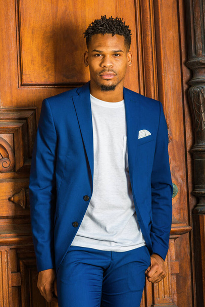 African American Businessman working in New York. Wearing blue suit, unbuttoned, white T shirt, black college student with little goatee, standing by vintage library doorway on campus, looking forward. - Photo, Image