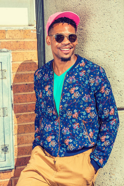 African American Man Street Fashion in New York, wearing blue patterned, collarless jacket, green V neck shirt, yellow brown pants, pink cap, sunglasses, standing against wall under sun, smiling - Fotó, kép