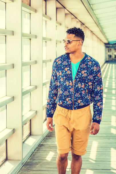 Moda casual de hombre joven afroamericano en Nueva York, con flores azules estampadas, chaqueta sin cuello, pantalones marrones amarillos, gafas de sol, caminando por la pasarela con pared de vidrio en el campus bajo el sol - Foto, imagen