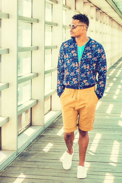 African American Man Moda casual a New York, indossando blu fantasia, giacca senza colletto, pantaloni gialli marroni, occhiali da sole, scarpe da ginnastica bianche, camminando su passerella con parete di vetro nel campus sotto il sole - Foto, immagini