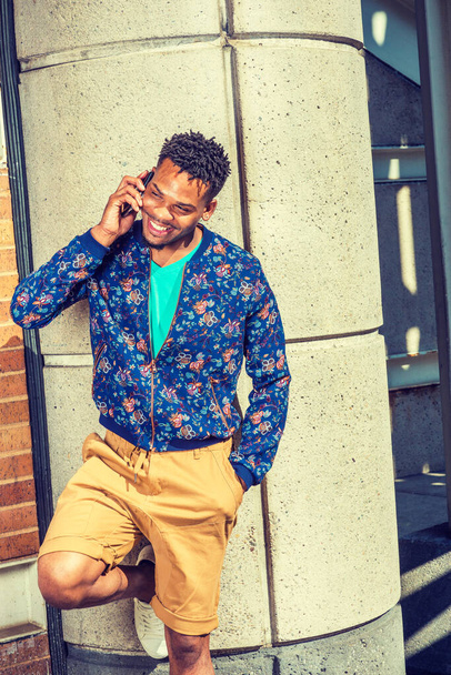 Fröhlicher Afroamerikaner, der in New York unterwegs ist, in blau gemusterter, kragenloser Jacke, gelbbraunen Shorts, in einer Straßenecke stehend, lächelnd, telefonierend. Moderne Straßenmode - Foto, Bild