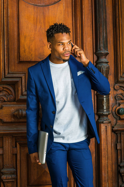 African American Businessman working in New York. Wearing blue suit, white T shirt, college student with little goatee, standing by vintage library doorway, holding laptop computer, talking on phone - Foto, Imagen