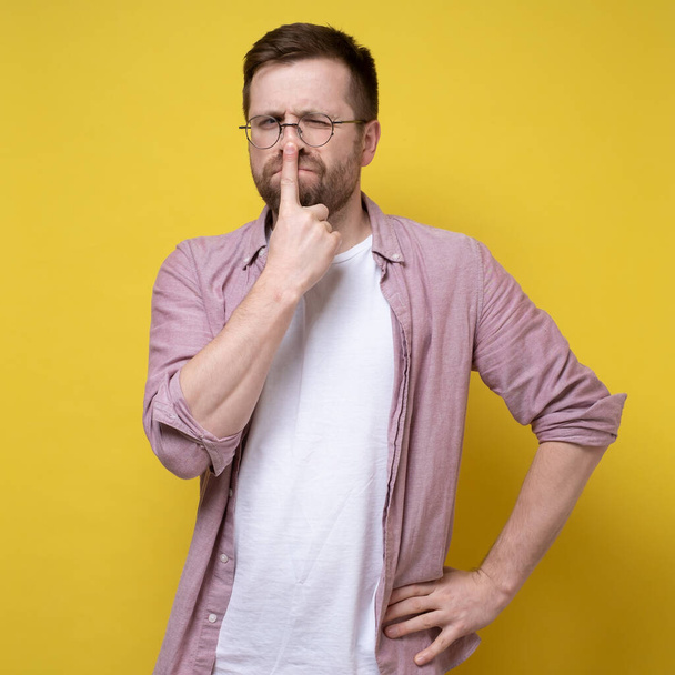 Young attractive man in glasses thought about something, he put finger to his nose and screwed up one eye. Studio, yellow background.  - Фото, изображение