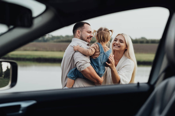 Vista a través de la ventana interior del coche, familia joven feliz con los niños abrazándose juntos y disfrutando de fin de semana fuera de la ciudad - Foto, Imagen