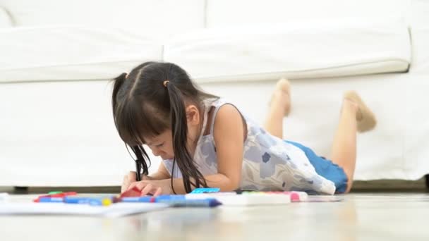 Милая очаровательная азиатская этническая девочка, держащая цветной рисунок ручкой и живопись, лежа на теплом полу, она развлекается и смеется. Концепция обучения и наслаждения творческим хобби, развития ребенка. - Кадры, видео