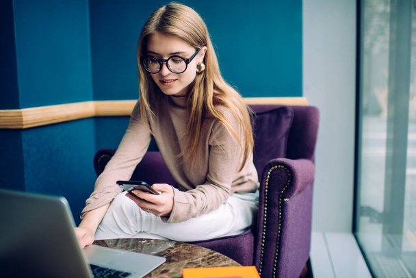 Jonge vrouwelijke student in casual trui en trendy bril zitten in moderne paarse fauteuil en surfen op internet op laptop tijdens het gebruik van smartphone - Foto, afbeelding
